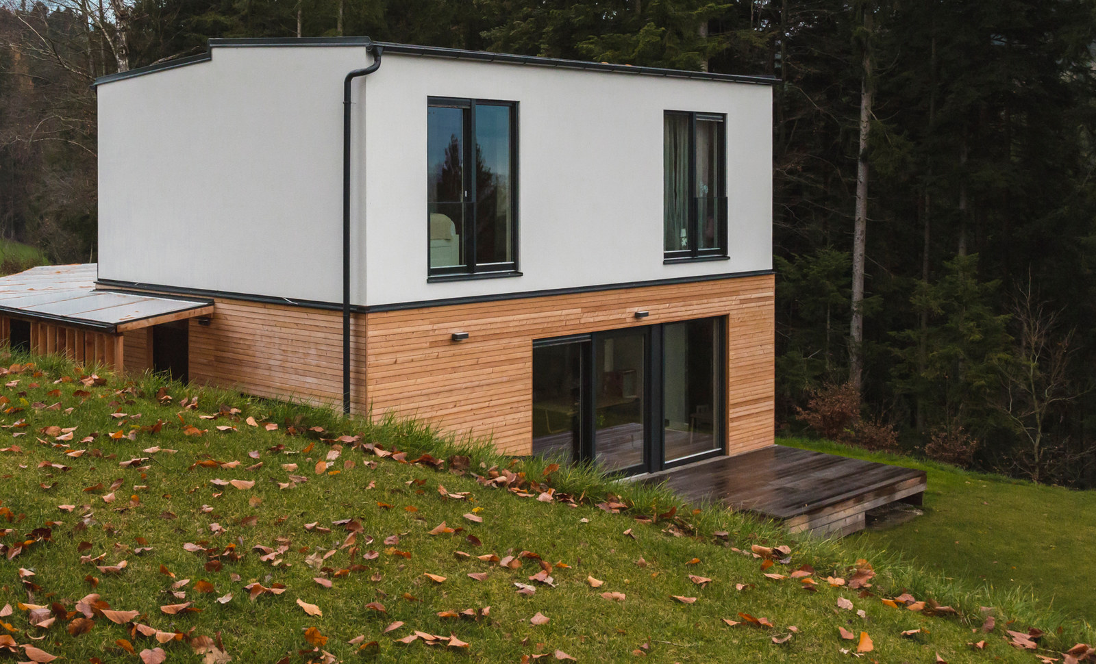 Zweigeschossiges COMMOD HOUSE am Hang, unteres Geschoss mit Holzfassade, Herbstlaub im Gras